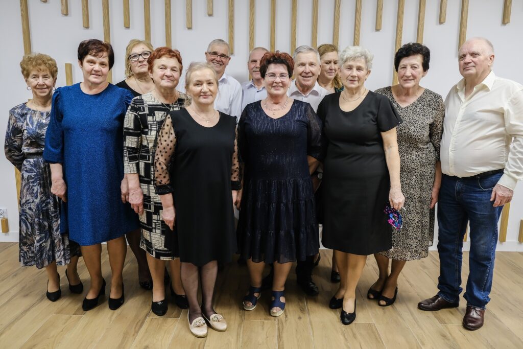Grupa kobiet i mężczyzn w wieku senioralnym ustawiona w kilku rzędach do wspólnej fotografii na tle białej ściany ozdobionej pionowymi lamelami